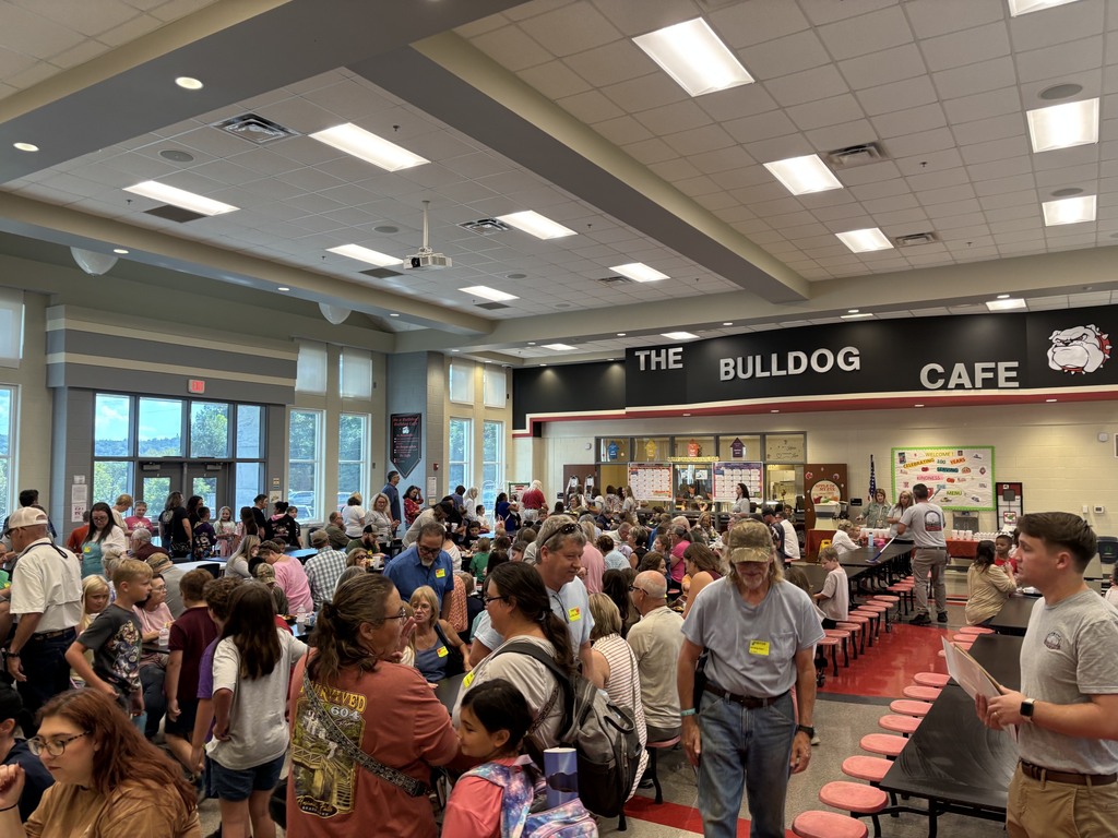 Crowd gathered inside a school cafeteria with a “THE BULLDOG CAFE” sign above serving counters, people in line near red flooring and tables, and posters on the walls.