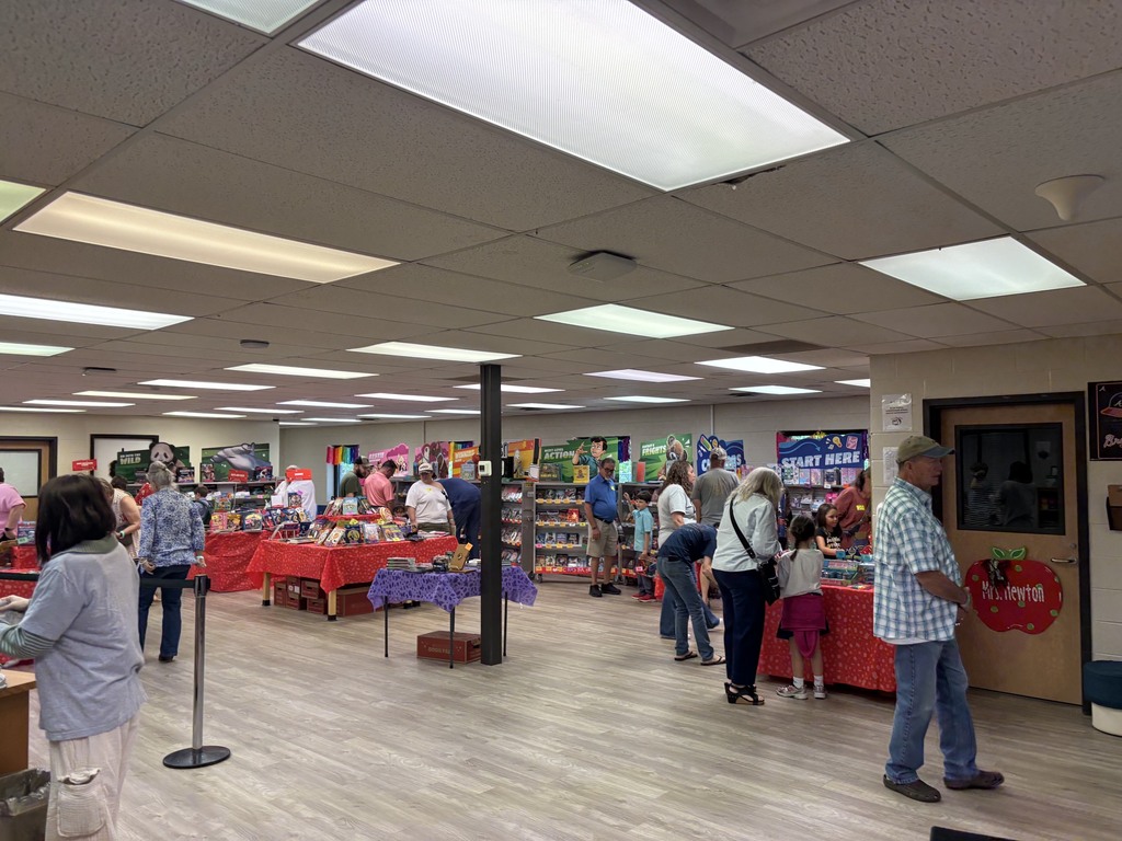 Busy indoor bookfair in a school Media Center, with people browsing tables covered in colorful toys and books also on displays under bright ceiling lights.