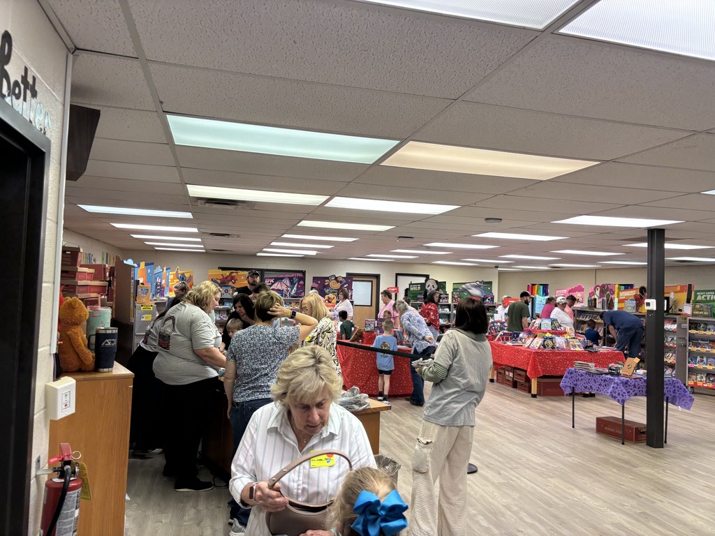 Busy indoor bookfair in a school Media Center, with people browsing tables covered in colorful toys and books also on displays under bright ceiling lights.