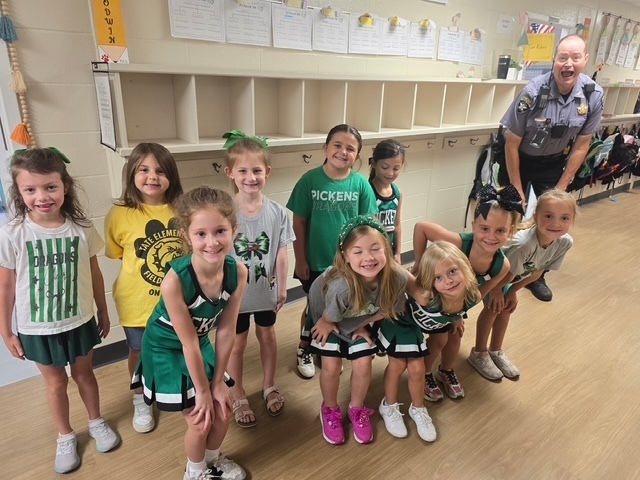 A group of children pose for a photo with an adult police officer standing behind them, smiling; kids wear green-and-white sports shirts and shorts, with cubbies in the background.