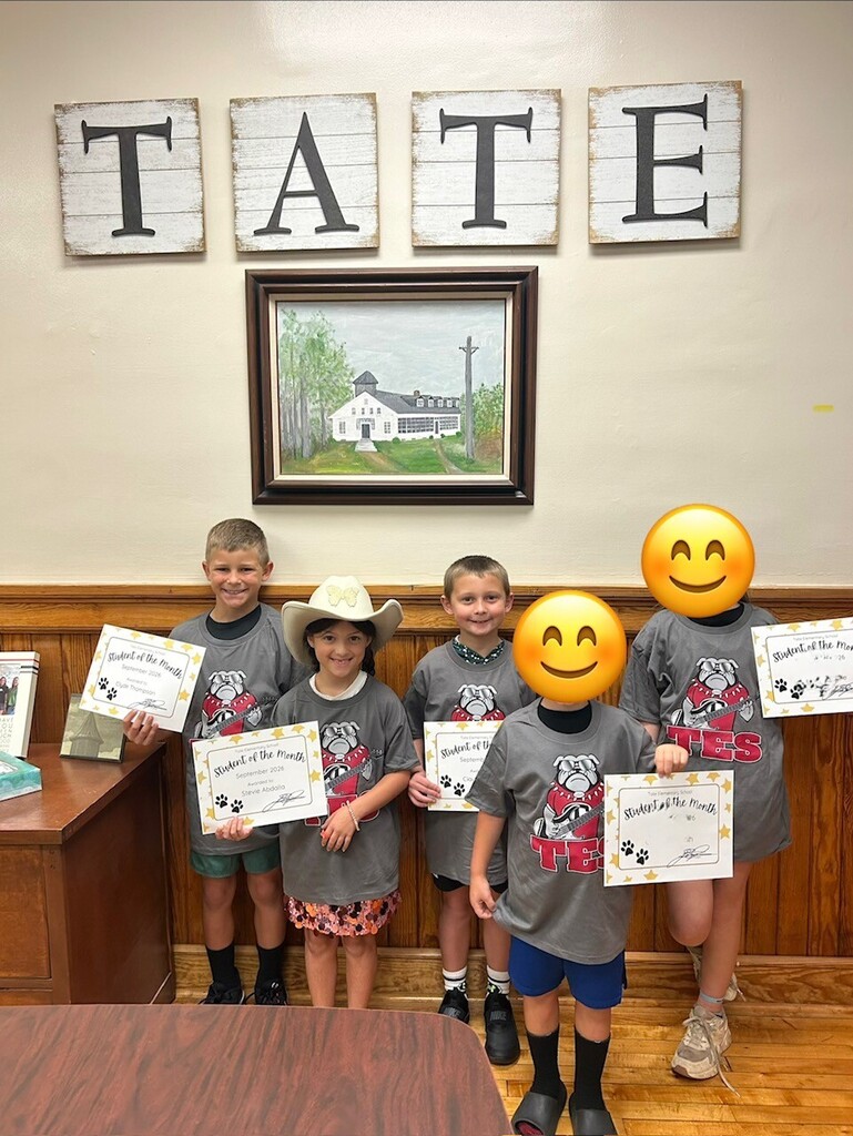 Five children stand indoors holding signed certificates, smiling toward the camera. Above them are wall letters spelling “TAITE” and a framed landscape picture; a wooden counter is in the foreground.