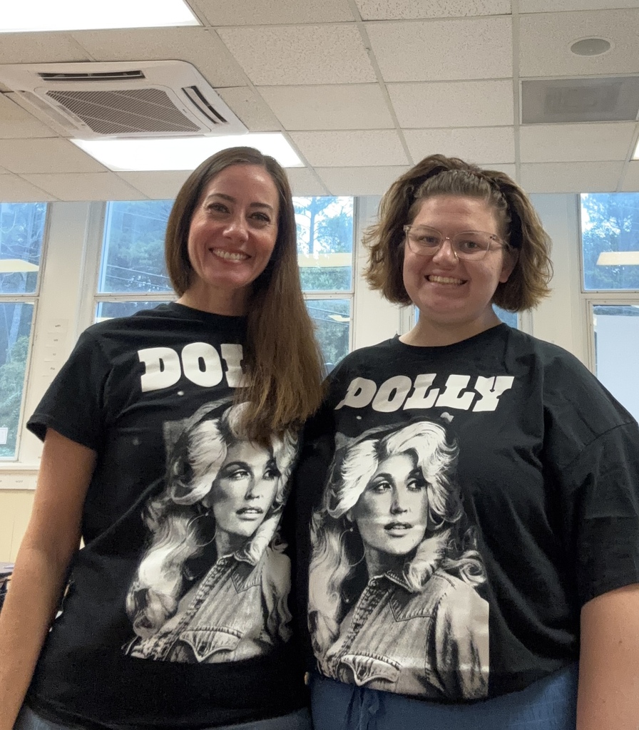 Two people smiling in an indoor office wearing matching black T-shirts with large grayscale portraits and the word “DOLLY” across the front.