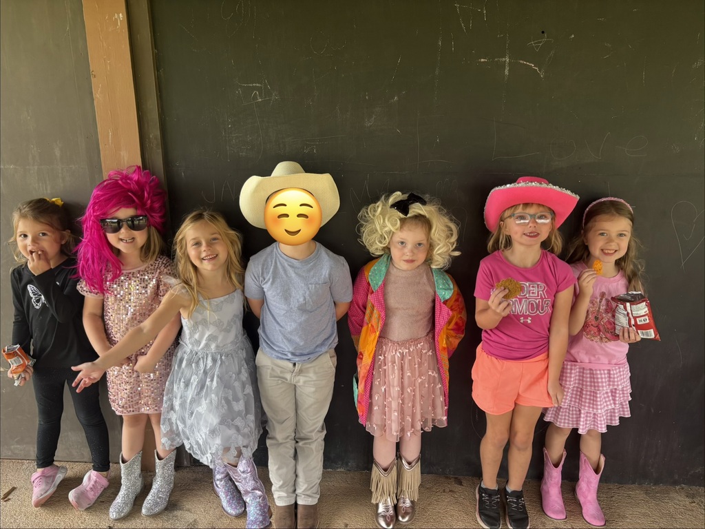 Seven children in colorful costumes pose together in front of a dark wall, wearing wigs and hats, with one child’s face covered by a yellow emoji icon.