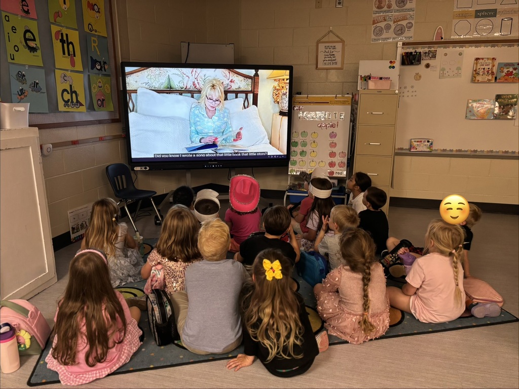 A group of children seated on a classroom floor watching a teacher on a TV screen, with posters on the walls and chairs and bins along the sides of the room.