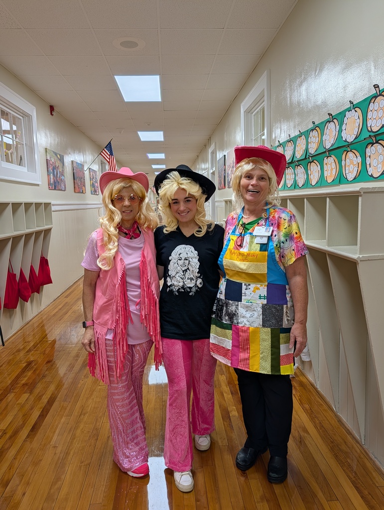 Three people pose together in a school hallway dressed in colorful costumes—pink outfit with hat, black shirt with white hair wig, and a rainbow apron—standing near bulletin boards and flags.