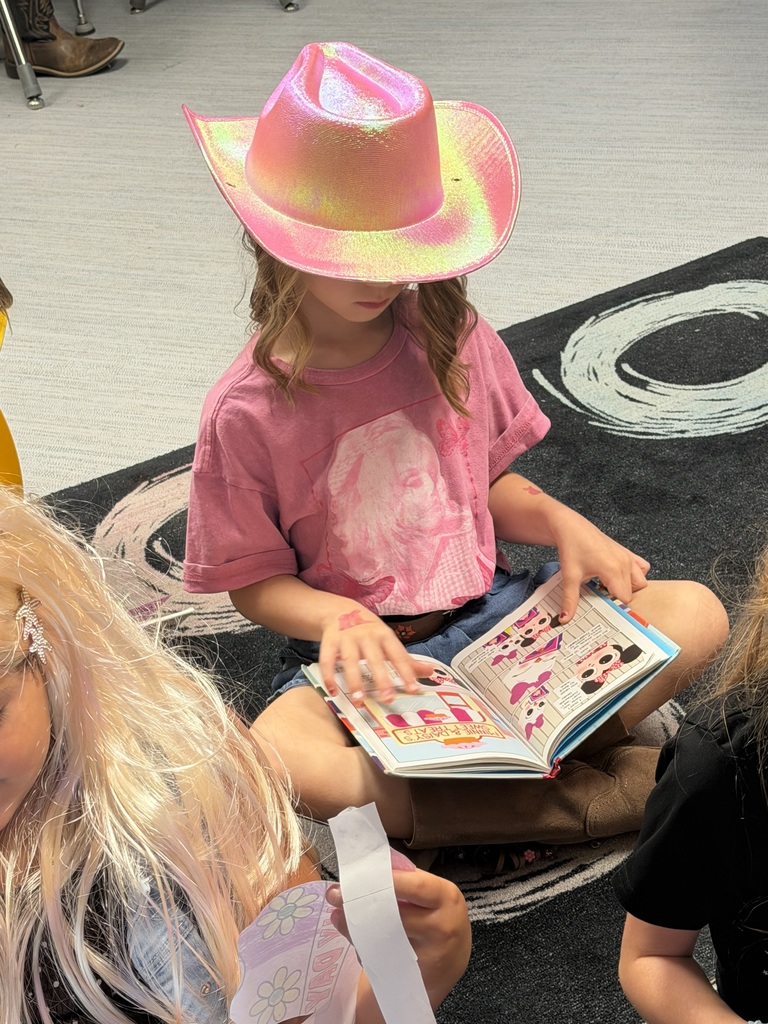 A child wearing a pink cowboy hat sits on a couch and reads an open picture book, while other people nearby watch in a bright indoor setting.