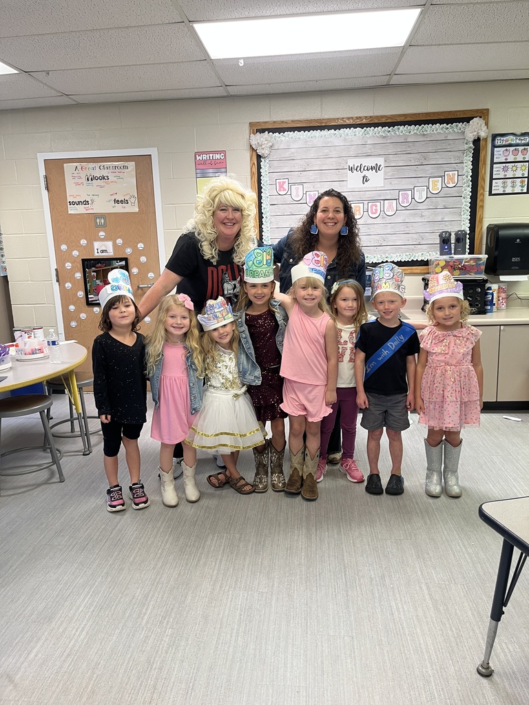 A teacher and a group of children stand together in a classroom wearing colorful hats and dressed in party outfits, smiling in front of a bulletin board and classroom supplies.