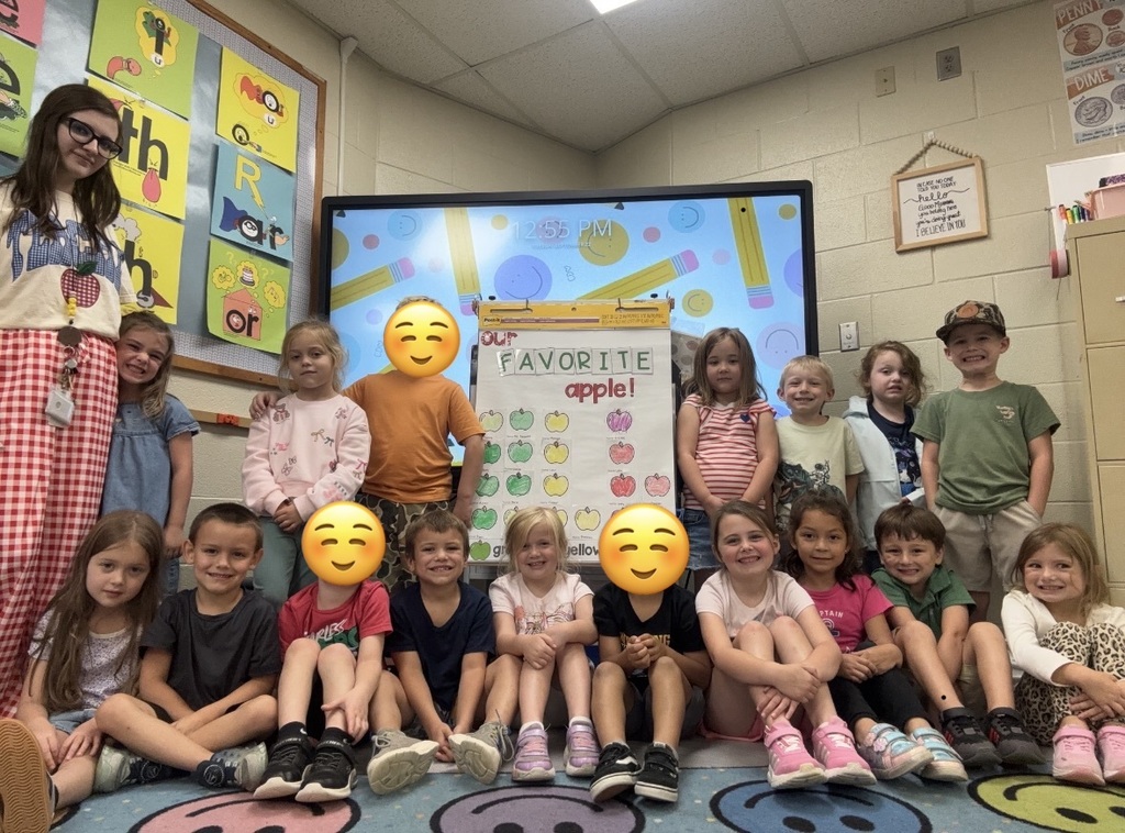 Group of children posing in a classroom in front of a colorful bulletin board and a TV screen that shows “Favorite Apple,” with several faces covered by yellow smiley icons.