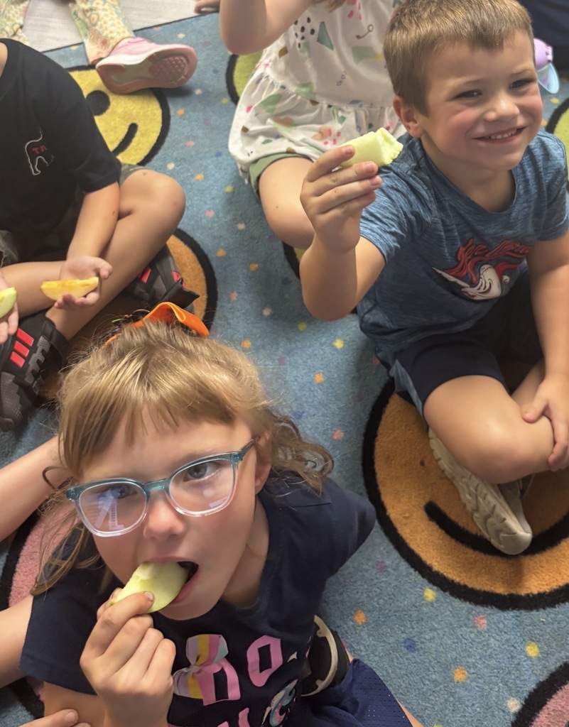 Children sitting on a blue carpet eating apples, including a smiling child in the foreground holding an apple and a girl with glasses eating an apple near the bottom left.