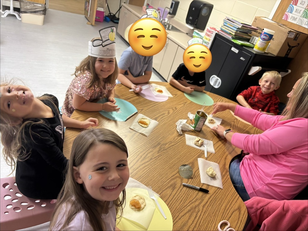 A group of children sitting around a wooden table during a meal or activity, with plates and food in front of them and several faces covered by yellow smiley icons in a classroom .