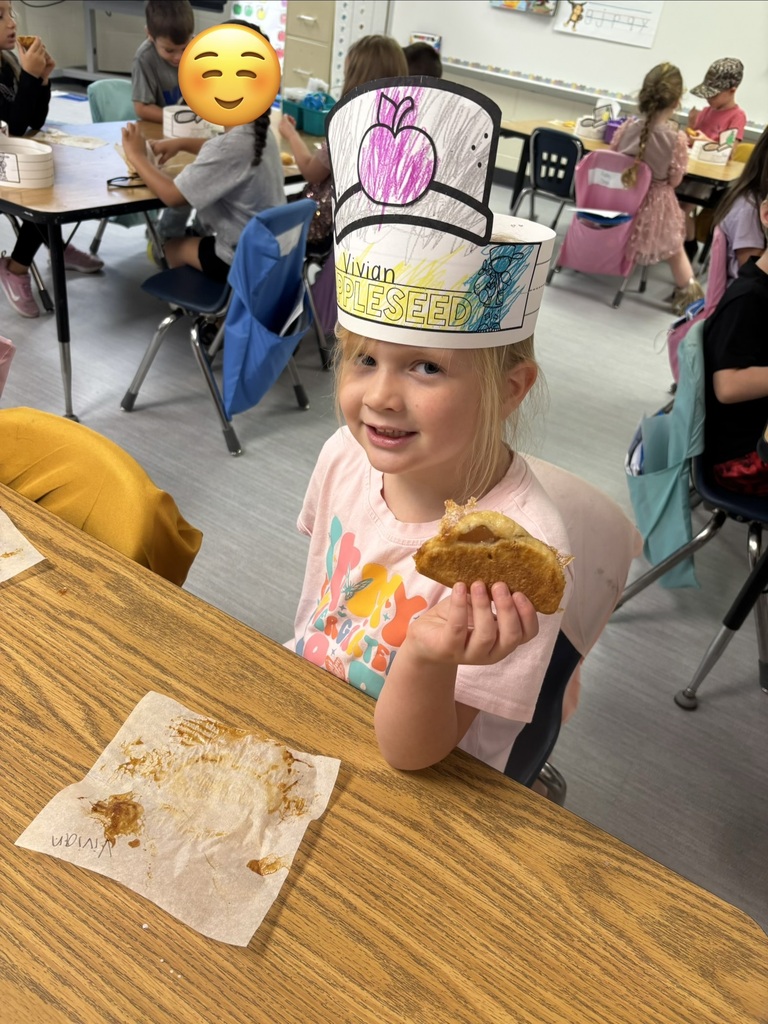 A smiling child wearing a handmade hat that reads “Living PLEASED” holds a small snack at a classroom table, while other kids sit in the background.