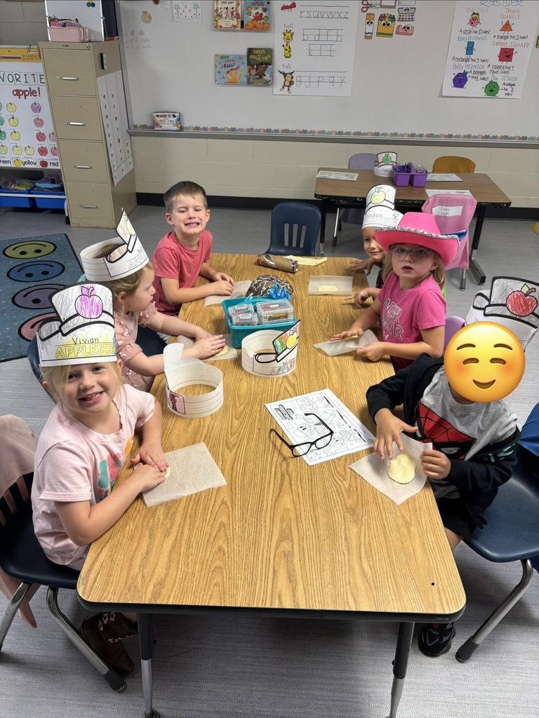 Children sit around a classroom table wearing paper crowns and hats, holding craft materials and drawings while smiling; a large smiley emoji appears at the bottom right.