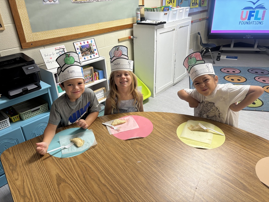 Three children wearing hats and smiling at a classroom table, each with a plate of food; colorful posters and play items surround them and a TV screen glows in the background.