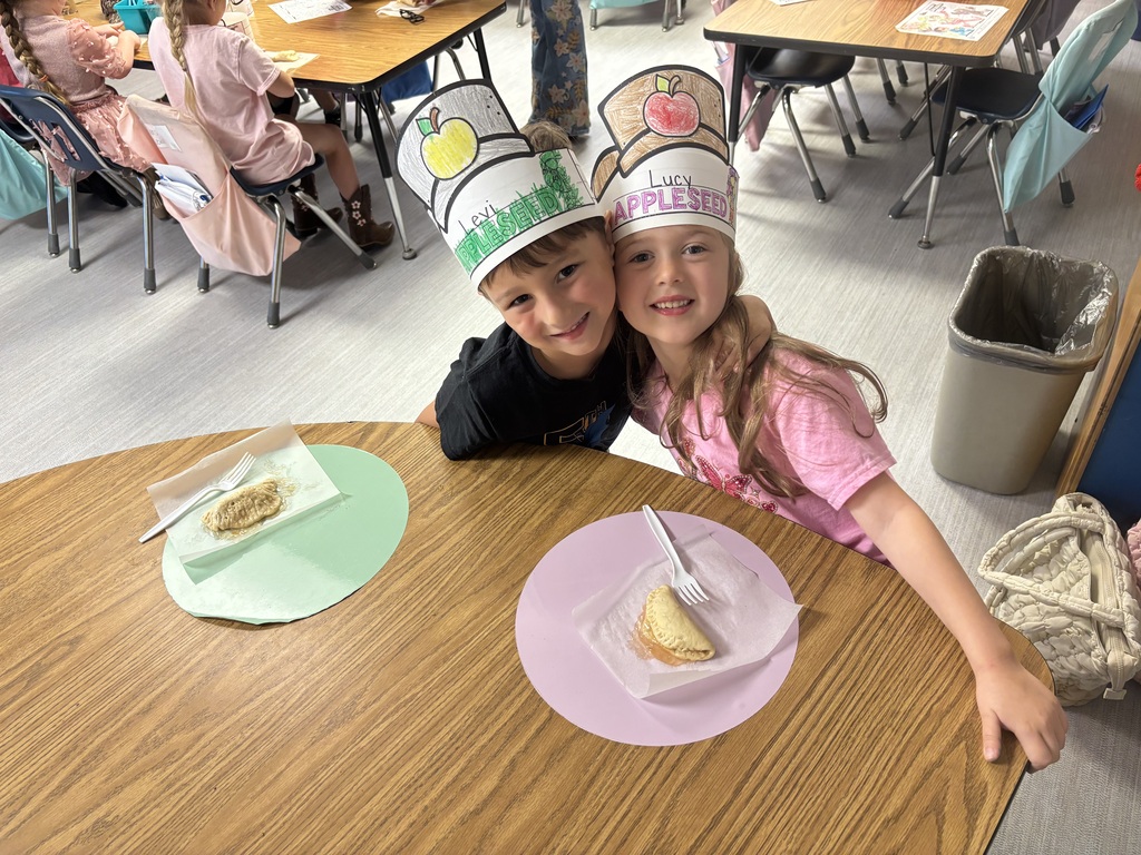 Two children sit at a classroom table wearing paper apple and fruit hats, smiling at the camera as plates with small snacks are placed in front of them.