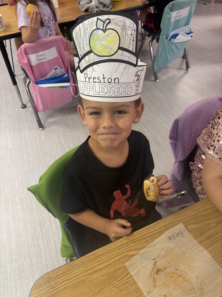 A child wearing a paper “Preston AppleSeed” crown with an apple drawing sits at a classroom table, holding a small snack while colorful chairs and name cards are visible behind.