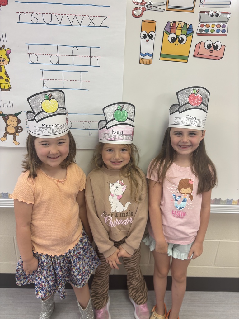 Three children stand side by side wearing paper chef hats decorated with crayons, smiling in front of a classroom wall with colorful drawings and letters.