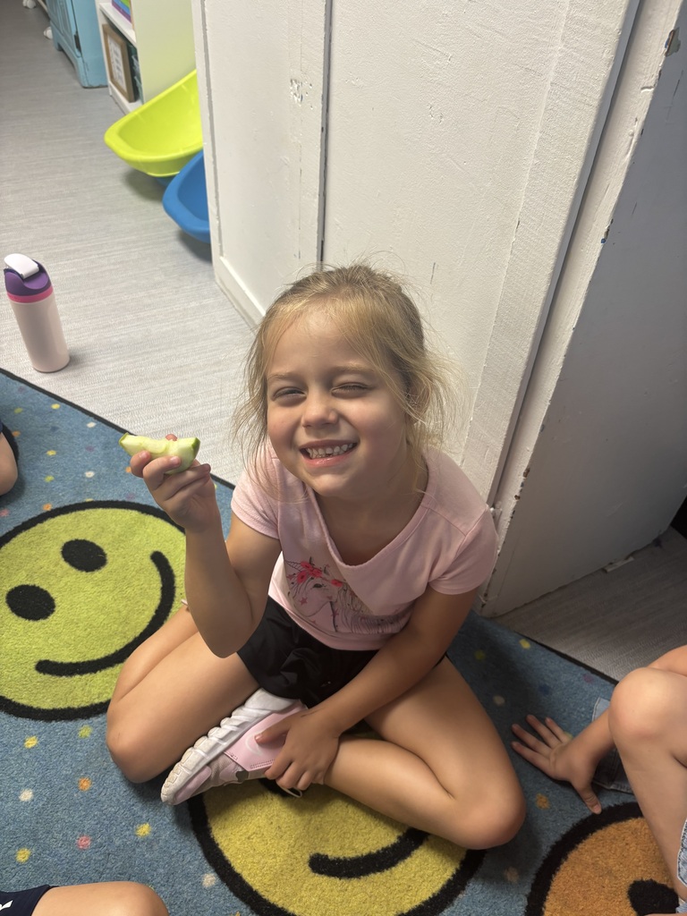 A smiling child sits cross-legged on a blue-and-yellow cartoon play mat near a wall, holding a small toy while another child’s hand reaches toward her.