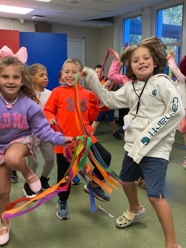 Children in a classroom wearing colorful hoodies hold long rainbow streamers while smiling and dancing near a window and classroom chairs.