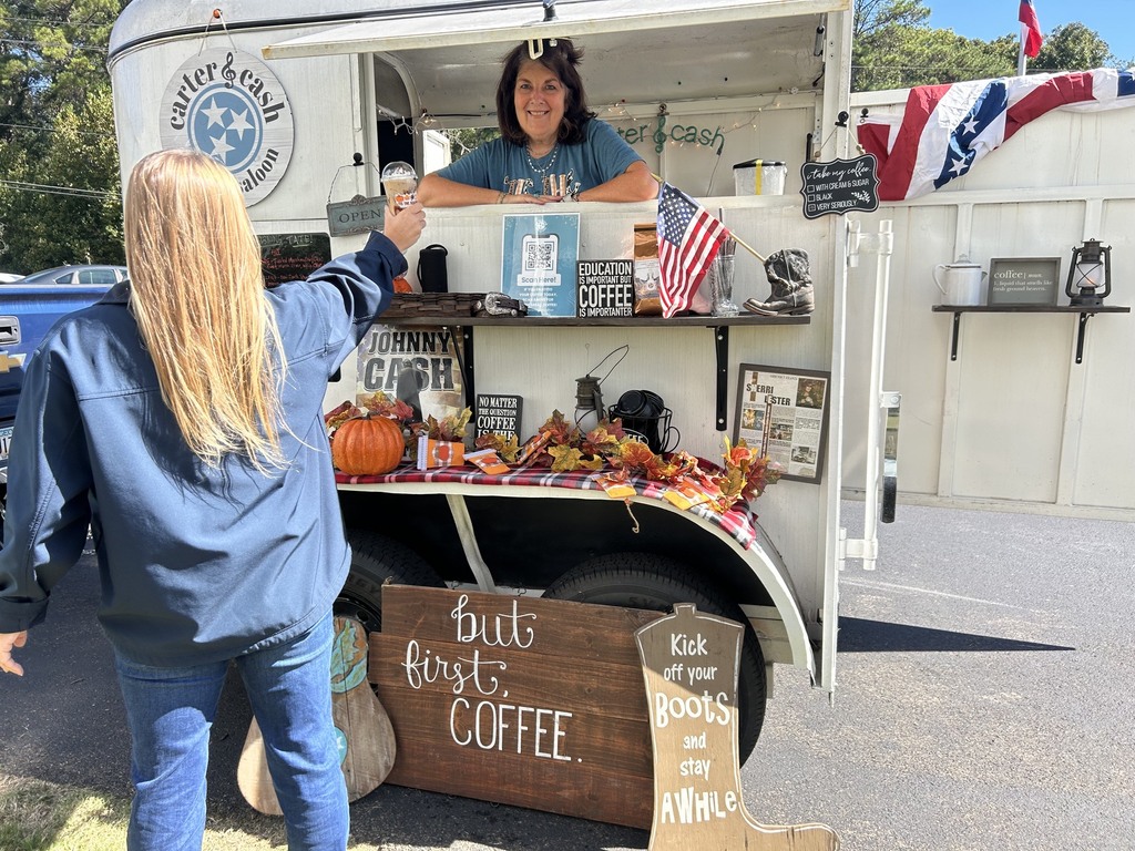 A woman serves coffee at an outdoor mobile coffee cart decorated for fall, with pumpkins and autumn garlands; a chalkboard sign reads “But first, COFFEE,” and a customer stands in front.