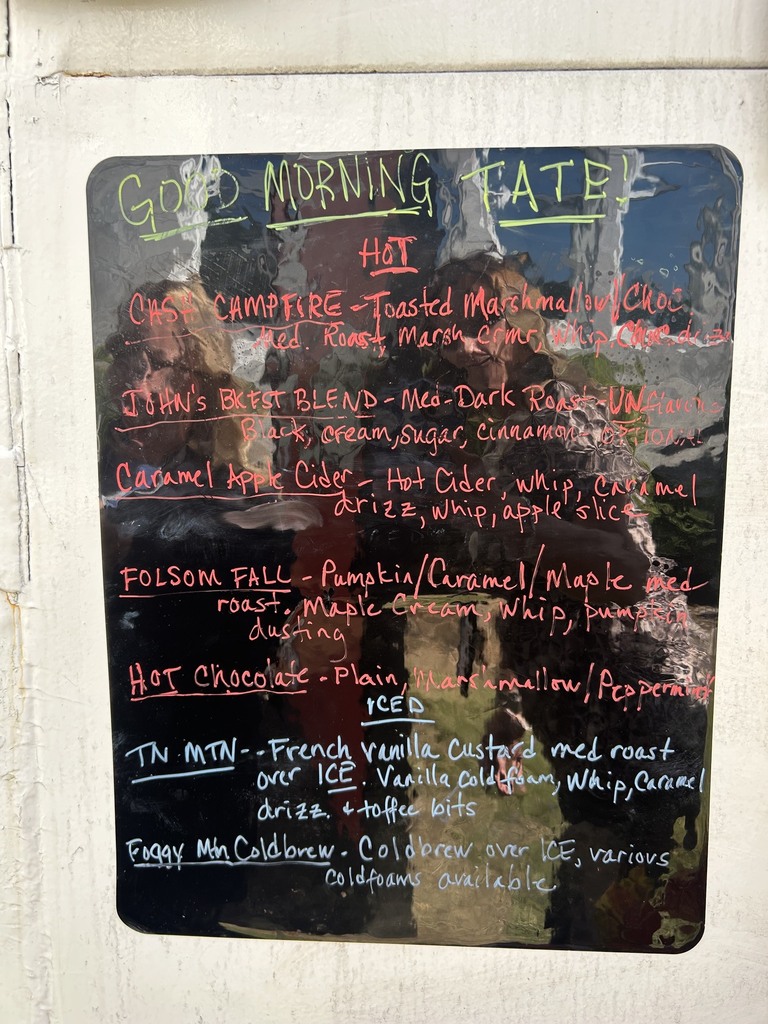 Blue chalkboard menu with colorful handwritten text reading “Good Morning Taters!” listing hot and cold drinks like campfire toasted marshmallow, caramel apple cider, Folsom Falo, hot chocolate, and French vanilla custard, with toppings.