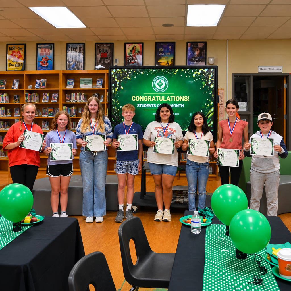 Seven PJHS students stand in the school media center holding Student of the Month certificates and wearing medals. A green “Congratulations! Students of the Month” display is behind them, with bookshelves and green balloons decorating the room.