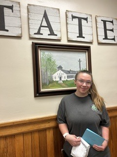 A person in a dark T-shirt stands indoors holding gifts and smiling, in front of a framed landscape painting and wall letters spelling “ T A T E”.