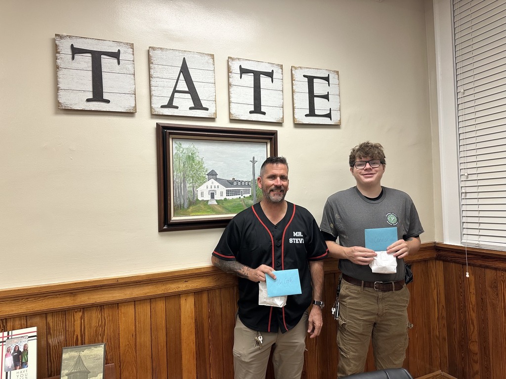 Two people standing holding light-blue envelopes, smiling in front of wall letters spelling “TATE” and a framed landscape painting.