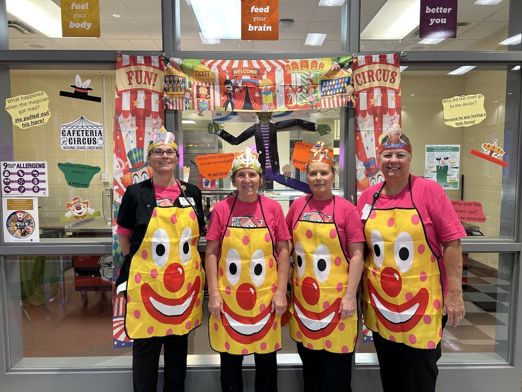 Four staff members wearing colorful clown aprons and paper party hats pose in front of a glass window decorated with a bright circus-themed banner and posters.