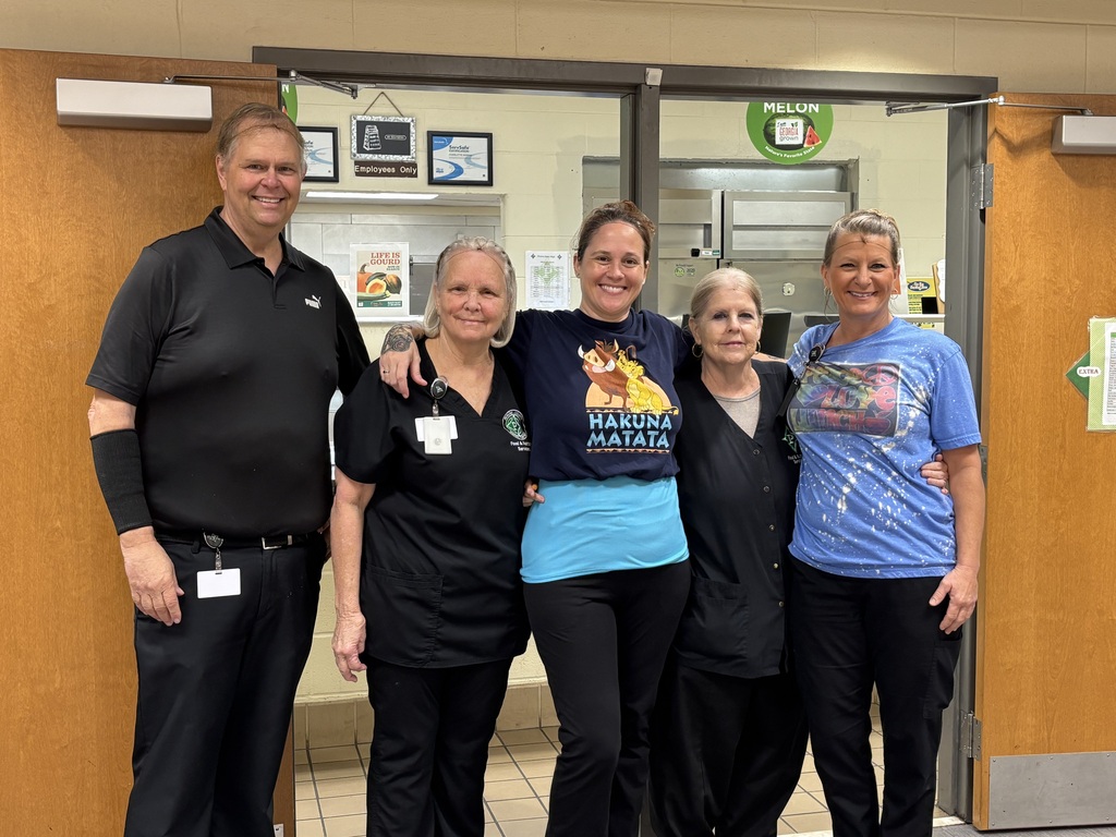 Five school cafeteria staff members stand together smiling in front of the kitchen serving area. They are dressed casually in black and blue shirts, with one staff member wearing a “Hakuna Matata” T-shirt.