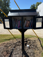 Black metal roadside book-sharing box with open top and several books visible inside, mounted on a post beside a gravel path and lawn.