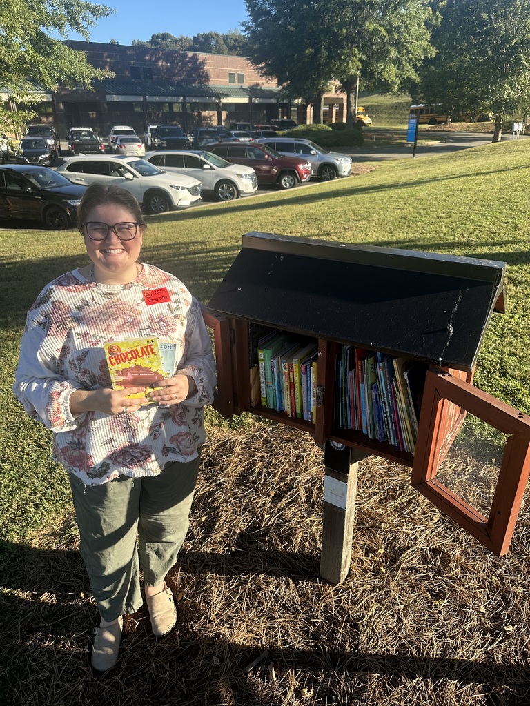 A person stands on a sunny lawn holding a book, smiling, beside a small outdoor bookshelf filled with colorful spines; a row of parked cars and buildings sit in the background.