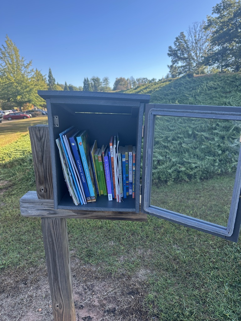 Open wooden book exchange box on a grassy hillside, with colorful books inside and a hinged glass panel on the right; trees and green hills under a clear blue sky in the background.