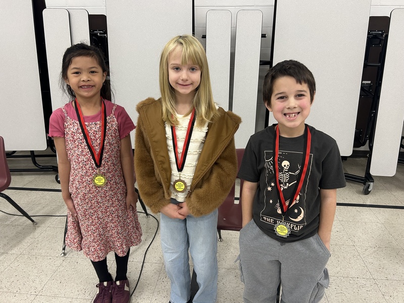 Three children stand in a row, smiling and wearing medals with red and black ribbons. They are in a room with white folding panels behind them.