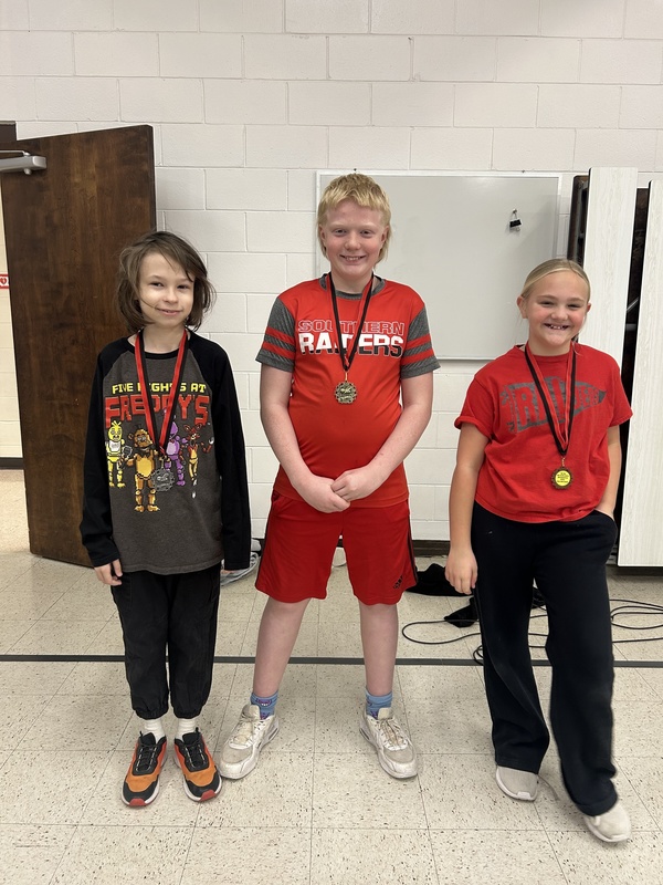 Three children stand smiling, each wearing a medal. The child on the left wears a graphic Freddy's shirt, the middle one wears a "Southern Raiders" uniform, and the child on the right wears a red shirt. A sense of pride is evident.