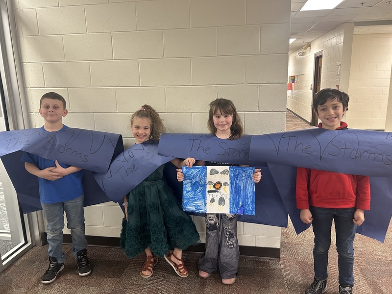 Four smiling children stand in a school hallway holding a large blue paper that reads "The Storms." One child holds a colorful drawing.
