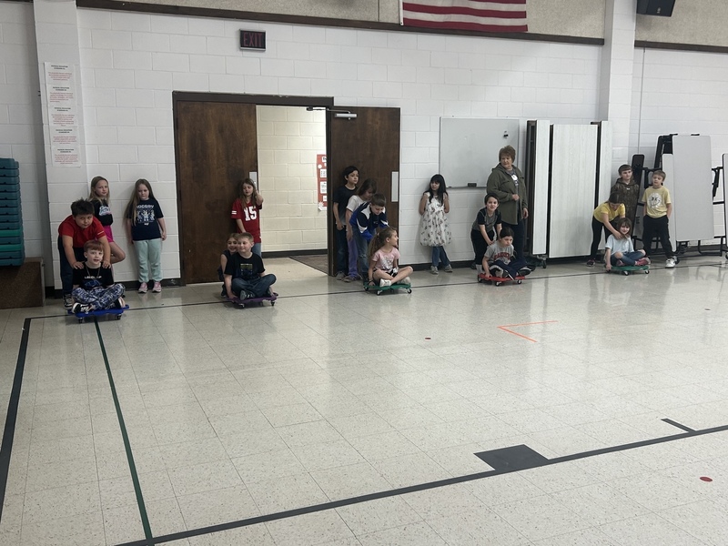 Children prepare for a scooter race in a gymnasium. They sit on colorful scooters, ready to start. A teacher oversees the playful atmosphere.