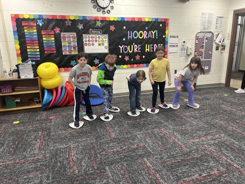 Five children stand in a classroom on paper plates, smiling and leaning forward. Behind them, a colorful bulletin board reads, "Hooray! You're Here!" The room has playful, vibrant decor.