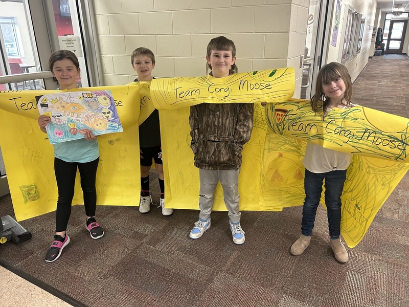 Four smiling children stand in a school hallway holding large, bright yellow posters with "Team Corg Moose" written on them. One child holds a colorful drawing. The atmosphere is cheerful and playful.
