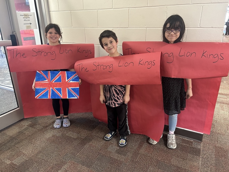 Three children smile while holding large red papers with "The Strong Lion Kings" written on them. One child holds a Union Jack poster.