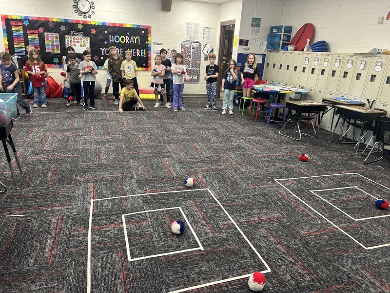 A classroom with children playing a game, aiming red, white, and blue balls at square targets on the floor. There's a vibrant, cheerful atmosphere.