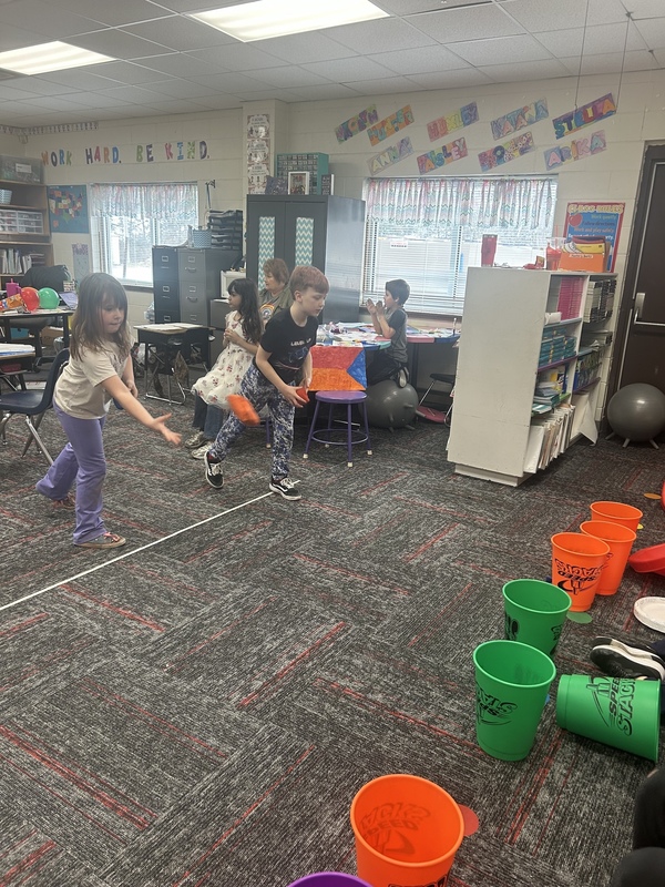 Children playing a bean bag toss game in a colorful classroom. Several kids aim at orange and green buckets while others watch. The atmosphere is playful.