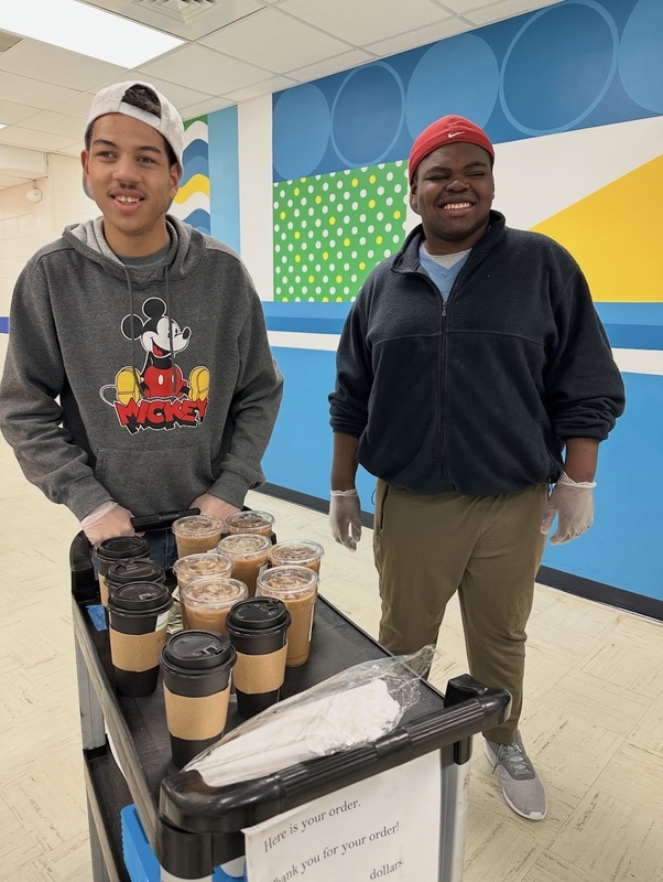 Two students from Palmer’s Pac smile while delivering Soaring Sips drinks on a cart, showing pride in their student-run coffee shop at MKL.