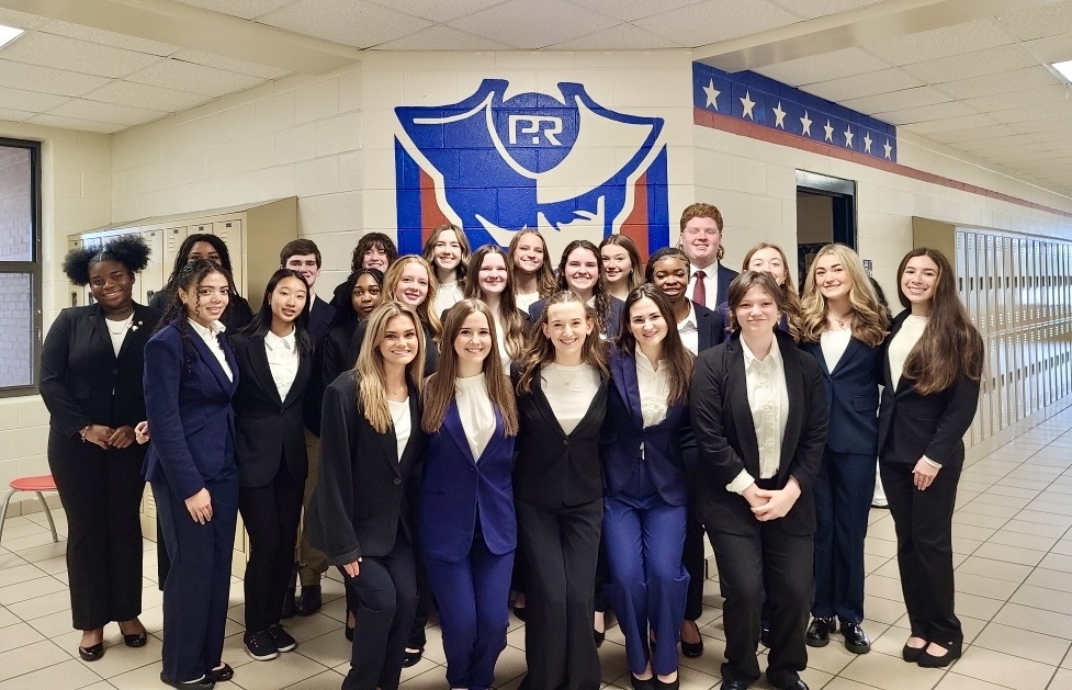 A large group of Pike Road High School HOSA students in professional blue and black suits pose together in a school hallway in front of a large blue and white "PR" shield logo. The students are smiling and celebrating their success at the Alabama HOSA State Leadership Conference.