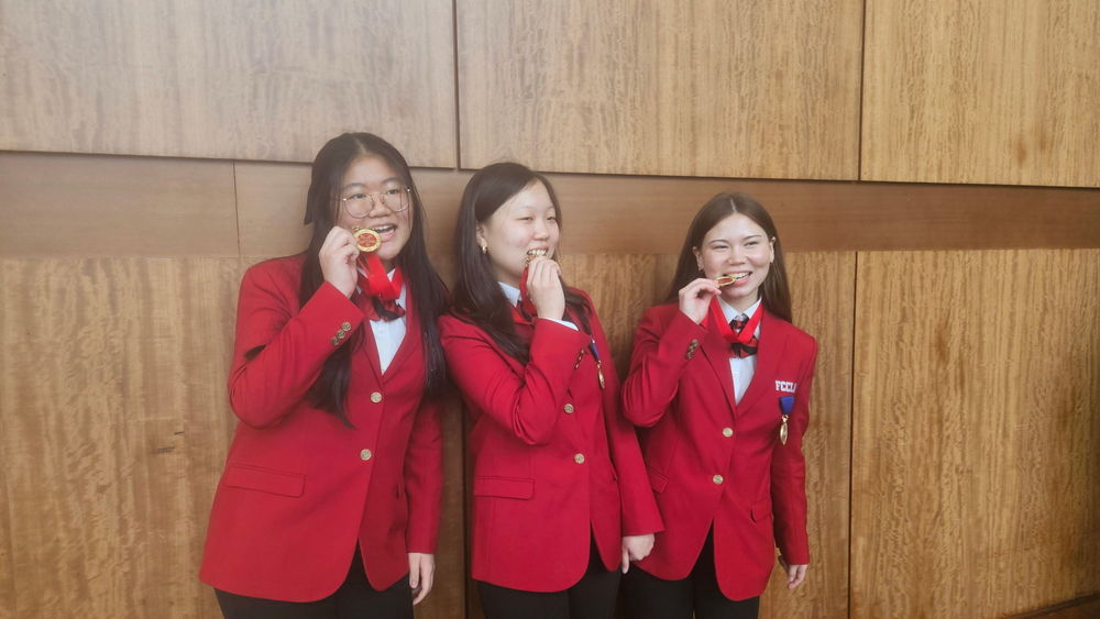 Three Pike Road Schools students wearing red FCCLA blazers smile and proudly pose with their medals. The student on the left holds a gold medal to her mouth, the student in the center smiles warmly, and the student on the right also poses with her medal against a wood-paneled wall background.