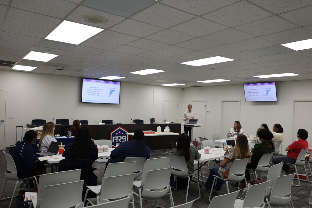 A professional development session held in a bright, modern conference room. A speaker stands at the front of the room near two large screens displaying presentation slides, while Pike Road Schools staff members sit at round tables, actively listening and engaging with the materials.