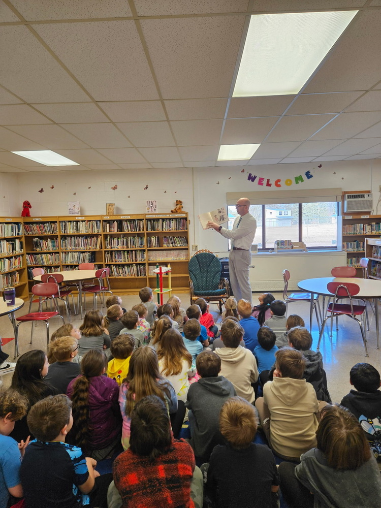 Superintendent Kowalczyk is standing in front of a group of students while reading a book.