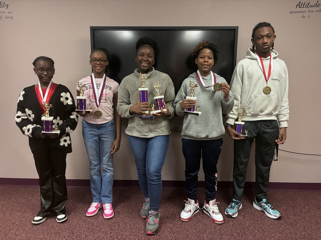 Five students stand together, holding trophies and medals, in front of a wall with a large screen.