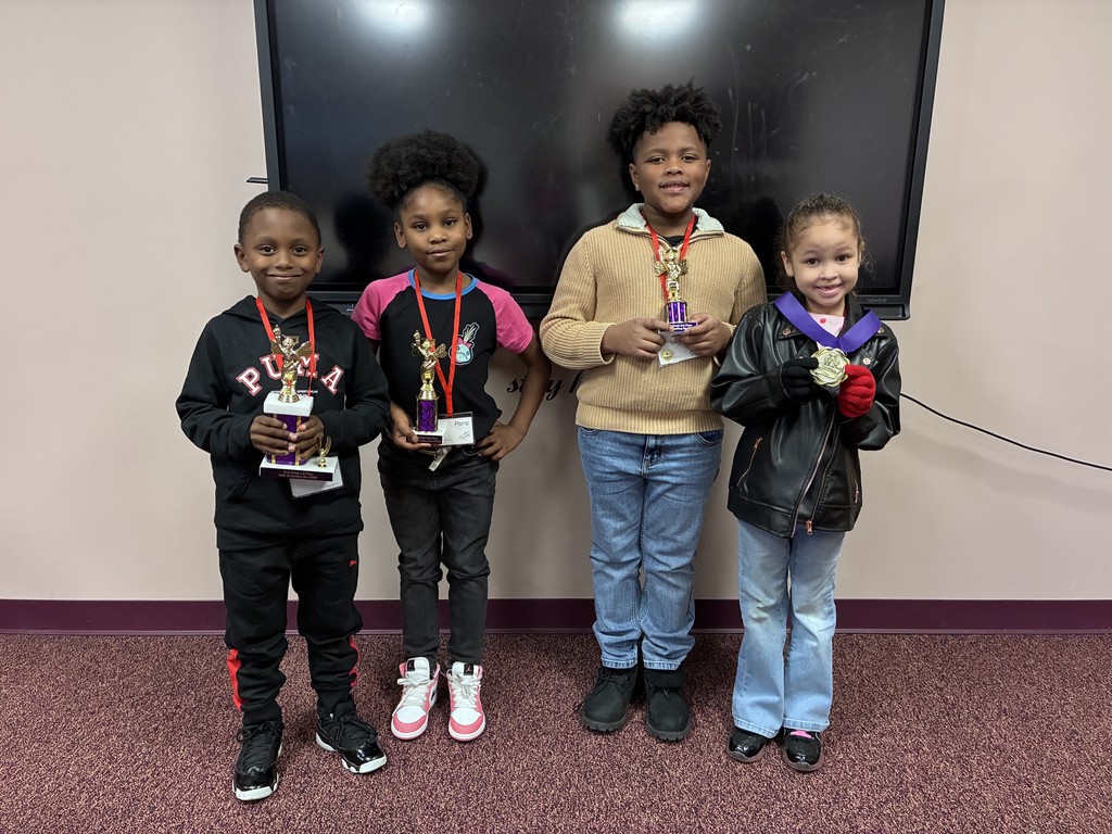 Four children stand in a row, holding medals. A large screen is on the wall behind them.