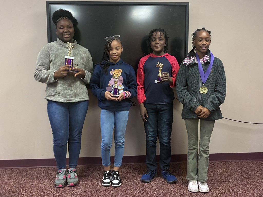 Four girls stand next to each other, holding trophies, in front of a blackboard.