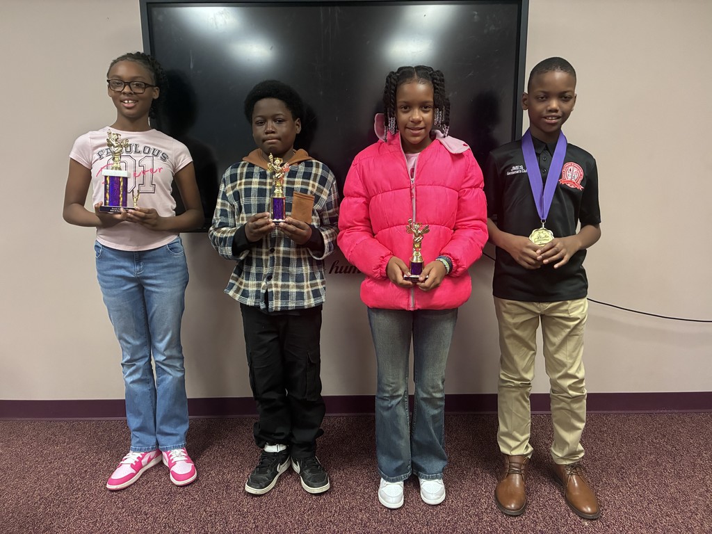 Four children stand together in a room, holding trophies. A large monitor is on the wall behind them.
